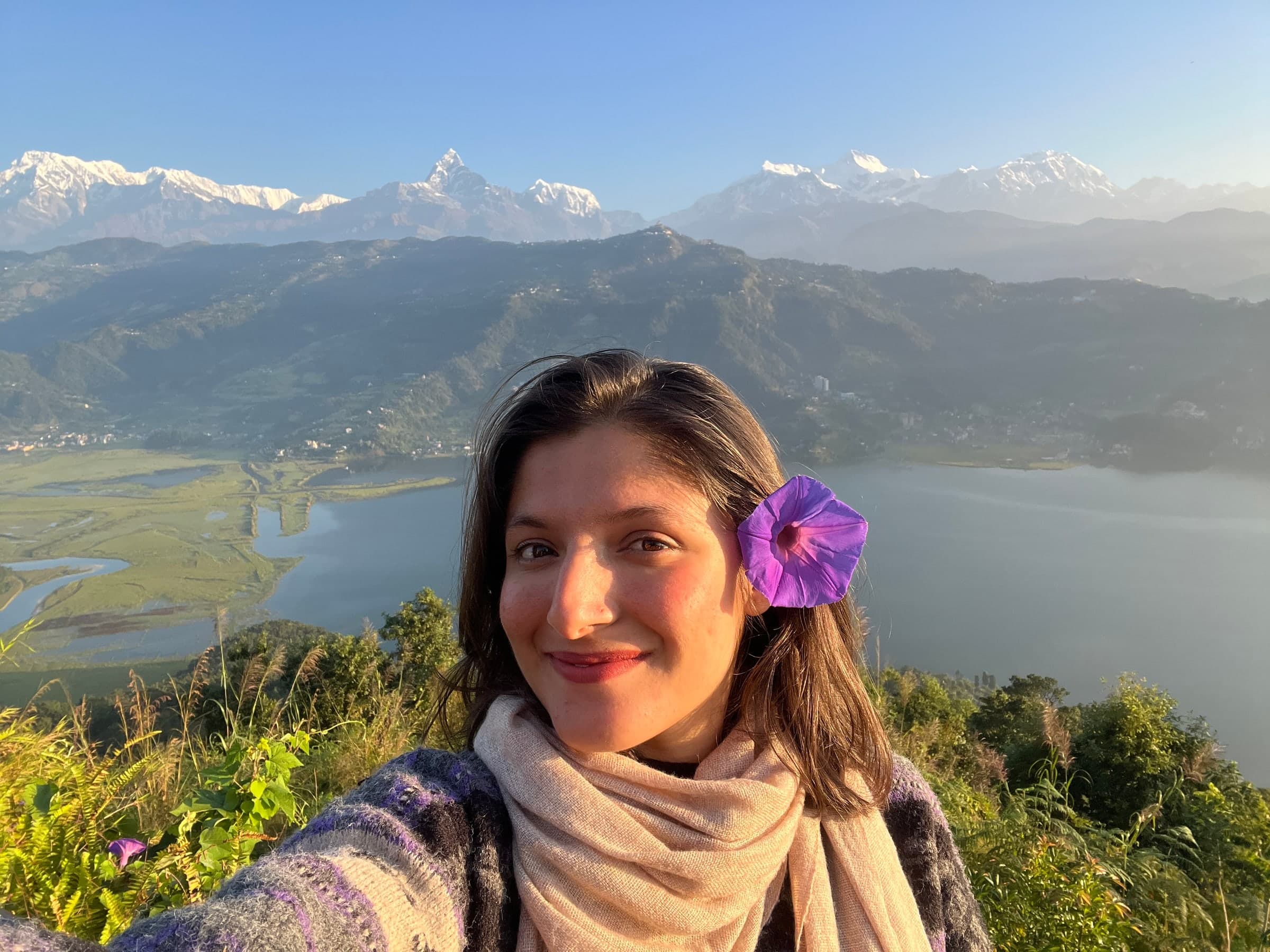 Pratiksha Paudel smiling, a purple flower in her hair, above a lake with snow mountains behind her
