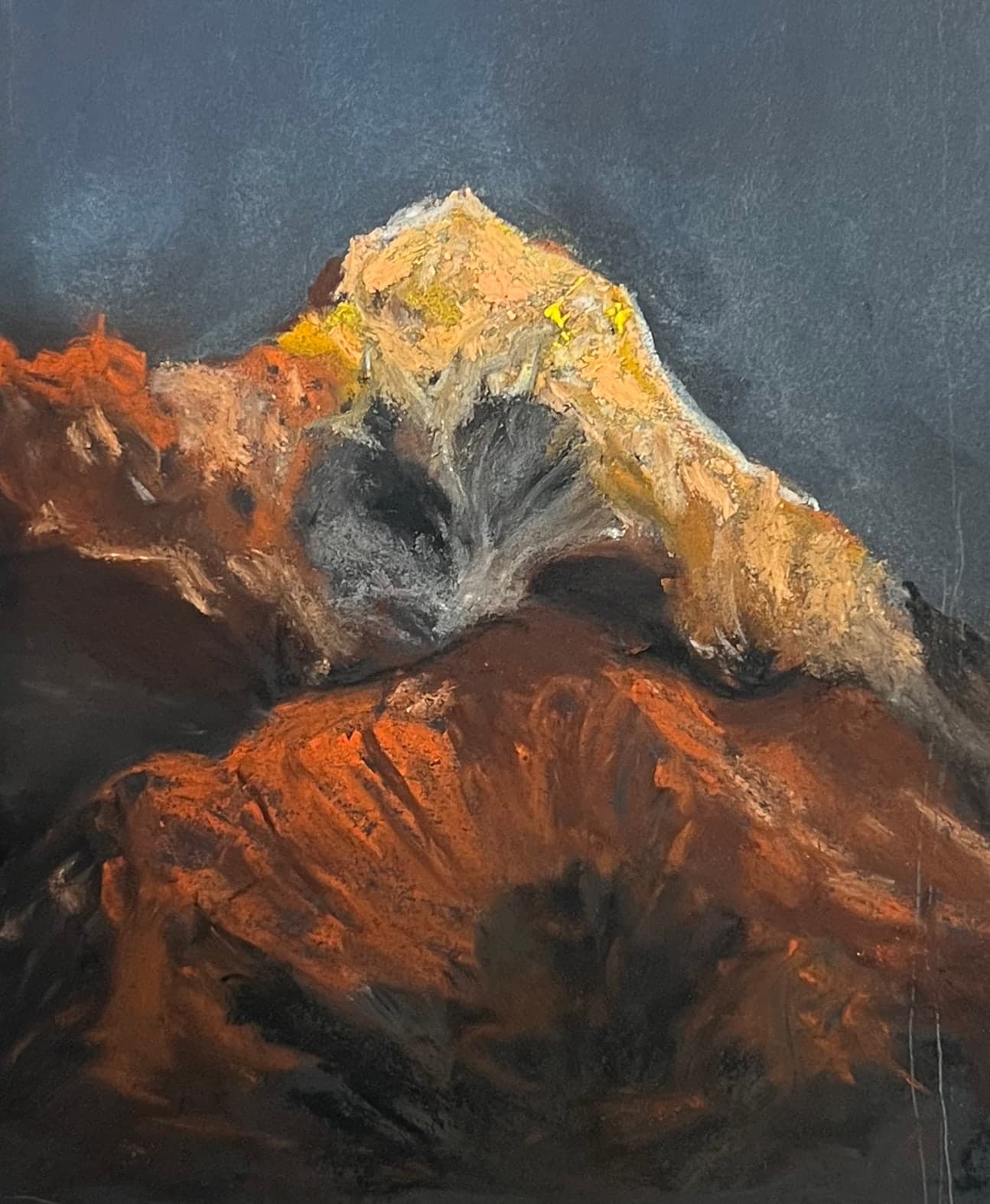 A mountain summit lit gold and orange by the last sun stands against a dark slate sky, its lower slopes burning rust red.
