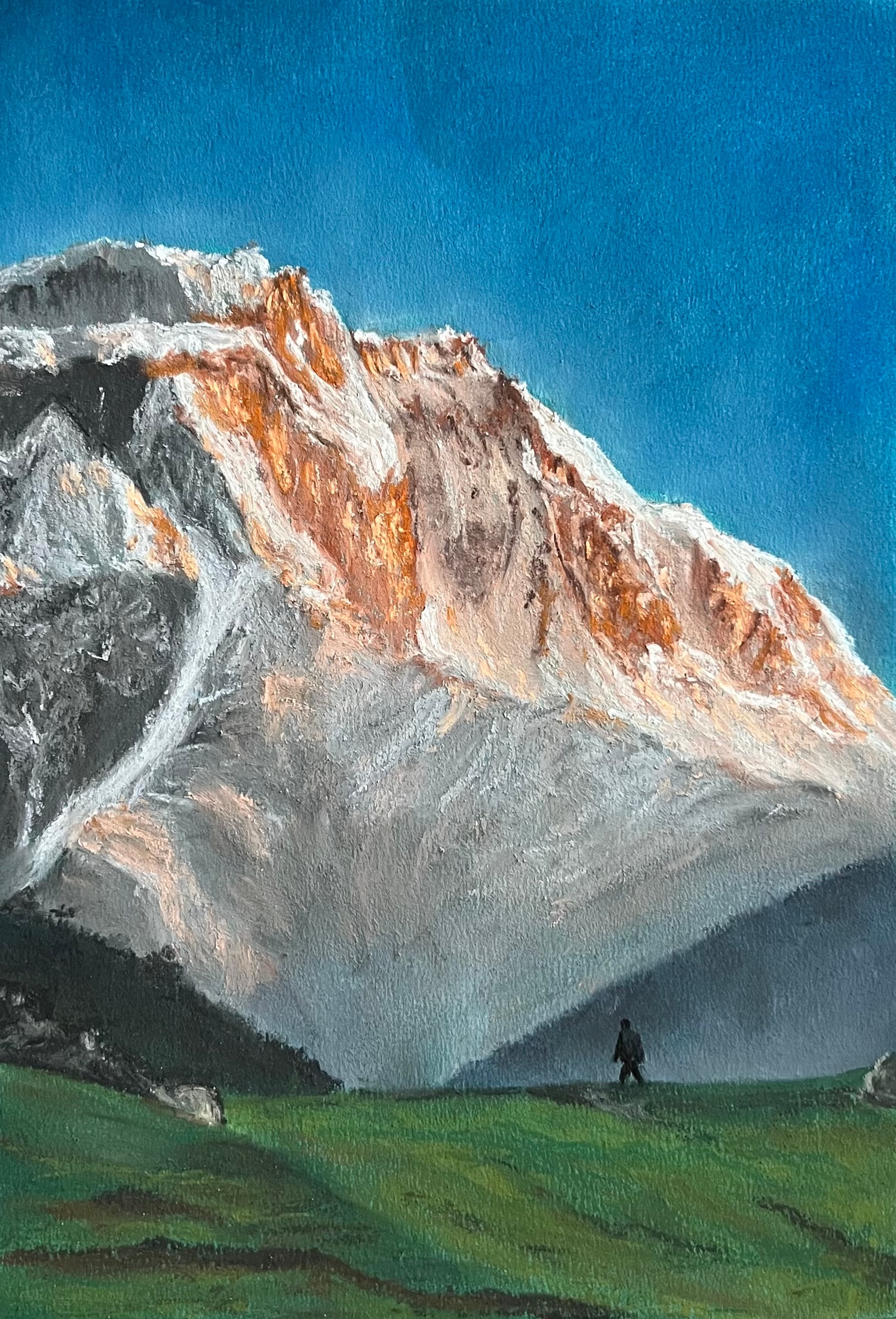 A lone walker crosses a green meadow at the foot of a huge rock-and-snow mountain face glowing peach in the sun, under a clear blue sky.